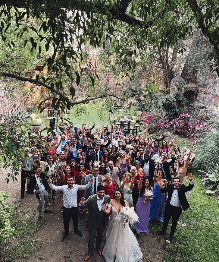 A newlywed couple, surrounded by a large group of smiling guests in the garden with their hands in the air.